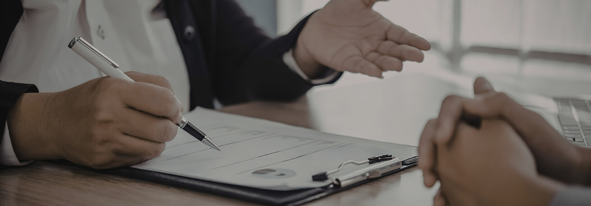Hands across a desk one person filling a form on a clipboard