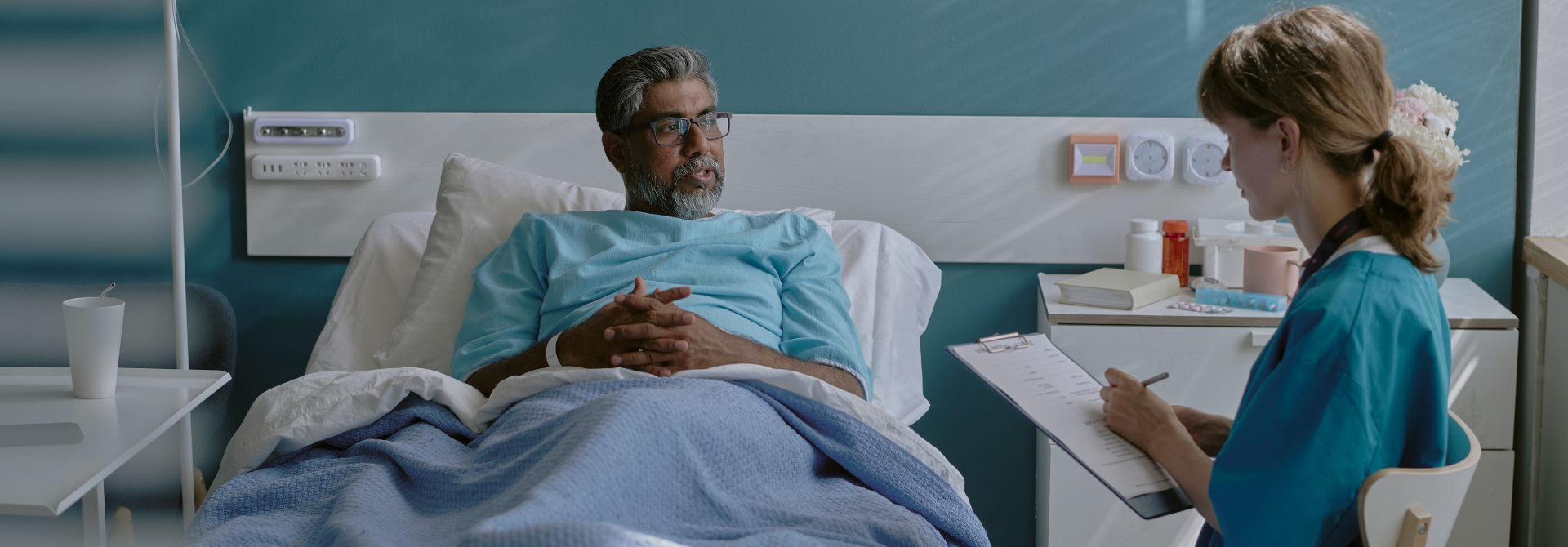 Man i a hospital bed speaking to a doctor filling in a form